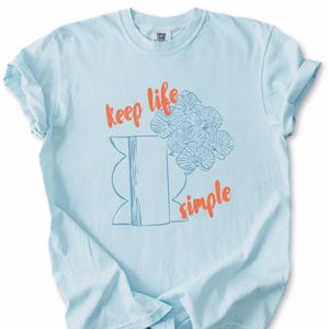 May include: Light blue t-shirt with rolled sleeves. The shirt features an orange text design that reads "keep life simple" with a blue illustration of flowers and a vase. The shirt is made of a soft material.