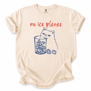 Op de afbeelding: Crèmekleurig T-shirt met een blauwe illustratie van een chagrijnige kat naast een glas ijsblokjes. De tekst "no ice please" staat in het rood boven de kat. Het shirt heeft opgerolde mouwen.
