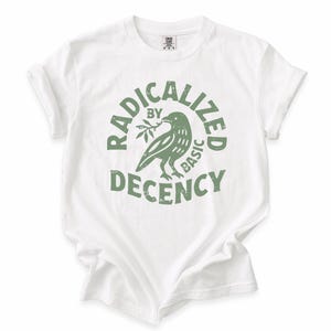 以下が含まれることがあります： 白いTシャツ、袖はロールアップされています。緑色のグラフィックで、小枝を持った鳥と「RADICALIZED BY BASIC DECENCY」という言葉が円形にデザインされています。このシャツは柔らかく快適な素材で作られています。