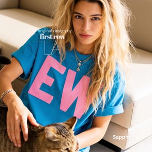 May include: A bright blue short-sleeved t-shirt featuring the large pink text "EW" on the front. The shirt includes the white printed text "original design by first row LA" near the neckline and the word "Sapphire" in the bottom corner.