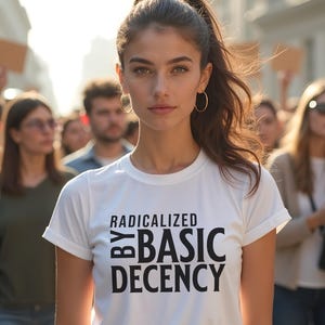 Puede incluir: Una camiseta blanca con el texto "RADICALIZED BY BASIC DECENCY" en negro. La camiseta la lleva una persona con pelo largo castaño y pendientes de aro dorados, de pie entre una multitud.