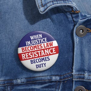 Pode incluir: Um botão redondo com o texto "WHEN INJUSTICE BECOMES LAW RESISTANCE BECOMES DUTY" em branco, vermelho e azul. O botão está preso a uma jaqueta jeans azul.