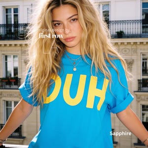 Op de afbeelding: Een felblauw T-shirt met korte mouwen en de grote gele tekst "DUH" op de borst gedrukt. Het shirt bevat de tekst "original design by first row LA" en het woord "Sapphire" in de rechterbenedenhoek.
