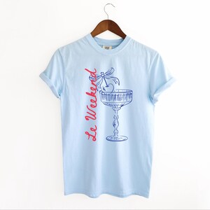 Chambray Light Blue T-shirt with the text Le Weekend and a Cherry Coquette Cocktail graphic.

Because what’s a good time without a little flair?

Perfect for brunch dates, rooftop soirées, or just channeling that effortlessly flirty energy. French twist, retro feel, full-on main character mood.
Weekend Shirt, Le Weekend, French T-Shirt, Weekending, Vintage Aesthetic, Coquette Bow Cherry, Cocktail Tee, y2k T-shirt, weekend vibes tee.