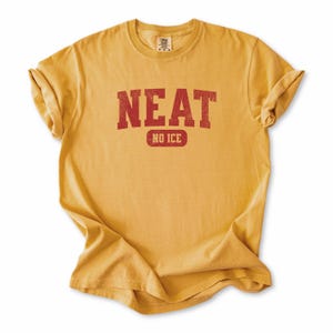 May include: Mustard yellow t-shirt with the word "NEAT" in red block letters above the words "NO ICE" in a red rectangle. The shirt is made of a soft material and has rolled-up sleeves.