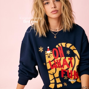 Puede incluir: Una sudadera azul marino de manga larga con el gráfico de un tigre amarillo que lleva un gorro de Papá Noel rojo y blanco. El texto "OH WHAT FUN" está impreso en letras rojas sobre el tigre, junto a pequeños copos de nieve dorados.