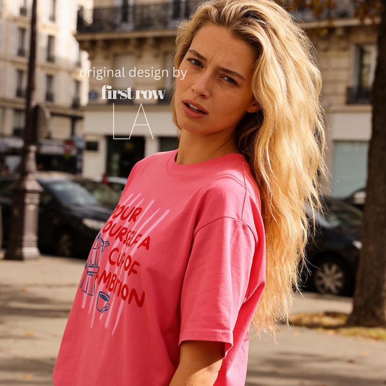 Puede incluir: Una camiseta de manga corta de color rosa brillante con el texto "Pour yourself a cup of ambition" en letras rojas, acompa&ntilde;ado de un gr&aacute;fico de una cafetera italiana y una taza de caf&eacute;. La prenda incluye el texto "original design by first row LA".