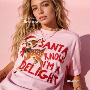 May include: A light pink short-sleeved t-shirt featuring a graphic of a fawn wearing a Santa hat and a red bow. The text reads "SANTA KNOWS I'M A DELIGHT" in bold red letters. The shirt also includes the text "original design by first row LA".