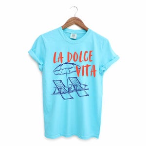 Lagoon Blue Tee with the text LA DOLCE VITA across the chest.
Two sun chairs. One umbrella. Zero worries. Italy -where the espresso hits hard, the gelato never melts, and responsibility? Never heard of her.
Endless summer, featuring a dreamy graphic of striped beach bliss. Perfect for sun-chasers, rule-breakers, and anyone who believes naps are a lifestyle.
La Dolce Vita shirt, Tomato Girl summer, Limoncello, spaghetti vibes. Retro foodie, Portofino, Amalfi Coast Shirt, Sardines Tee
