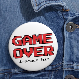 Op de afbeelding: Een witte button met de woorden "GAME OVER" in rode, gepixelde letters. Daaronder staat de tekst "impeach him" in een kleinere, zwarte letter. De button is geplaatst tegen een blauwe denim achtergrond.
