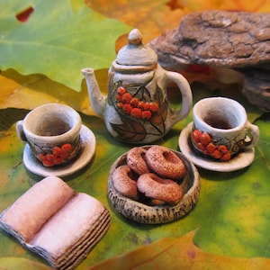 Puede incluir: Juego de té de cerámica en miniatura con tetera, dos tazas y platillos, un cuenco de rosquillas y un libro. La tetera y las tazas están decoradas con bayas rojas y hojas verdes. El juego está dispuesto sobre hojas de otoño.