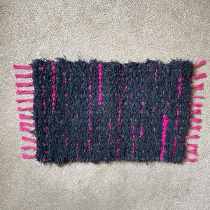 May include: A rectangular woven mat featuring a dark charcoal grey shaggy texture interspersed with vertical stripes of vibrant hot pink yarn. The edges of the mat are finished with short, knotted hot pink fringe tassels on both sides.