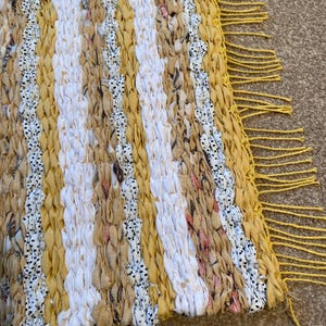 May include: A yellow and white woven rug with a fringe border. The rug is made of strips of fabric woven together to create a striped pattern.