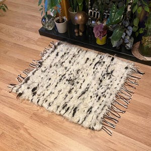 May include: A rectangular, off-white rug with a shaggy texture and black speckles. The rug has a fringe of black and white threads along the edges. The rug is placed on a wooden floor.