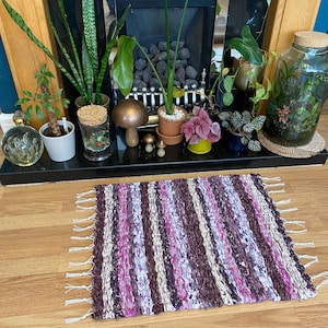 May include: A handwoven rug with a striped pattern in shades of purple, pink, and white. The rug has fringe on the ends and is sitting on a wooden floor.