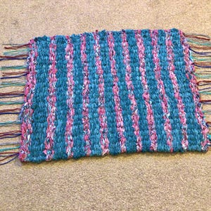 May include: A handwoven rug with pink and teal stripes and fringe. The rug is made from recycled materials and has a rustic, handmade look.