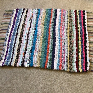 May include: A colourful, handwoven rug with a striped pattern and fringe on the edges. The rug is made from recycled materials and has a variety of colours, including blue, green, red, and white.