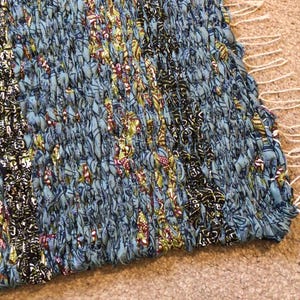 May include: A blue and gold woven rug with a fringe edge. The rug has a geometric pattern and is made from recycled materials.