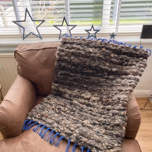 May include: A textured, brown and grey throw blanket with blue fringe is draped over a brown armchair. The blanket has a thick, nubby texture. The fringe is a bright blue colour. The blanket appears to be handmade.