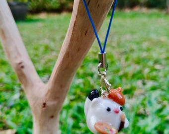 May include: A white ceramic charm of a cat wearing a brown beret and holding a paintbrush and palette. The charm is attached to a blue string and a silver clasp.