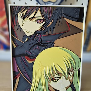 Op de afbeelding: Een ingelijst anime-kunstwerk met twee personages met gedetailleerde gezichtskenmerken en een gelaagd ontwerp. Het kunstwerk toont een mannelijk personage met donker haar en een vrouwelijk personage met blond haar. De lijst is wit, op een houten oppervlak.