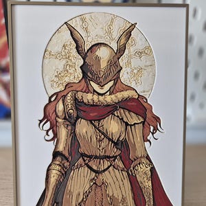 Op de afbeelding: Ingelijste illustratie van een personage in gouden harnas, met een rode cape en een gevleugelde helm. Het kunstwerk staat tegen een gestructureerde, cirkelvormige achtergrond. De lijst is neutraal van kleur en het kunstwerk staat op een standaard.
