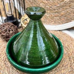 May include: A glossy, emerald green tagine pot with a conical lid and a wide, shallow base. The traditional Moroccan cooking vessel is made of ceramic and sits on a woven placemat. The tagine's lid has a rounded top with a small opening.