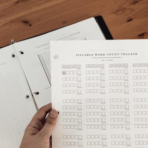 Może przedstawiać: Biała kartka zatytułowana "Fillable Word Count Tracker" jest trzymana przed segregator. Tracker ma rzędy kwadrat&oacute;w do śledzenia liczby sł&oacute;w. Segregator jest otwarty na stronie z siatką. Tło to drewniana powierzchnia.