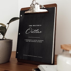 Puede incluir: Un portapapeles de madera con un clip plateado que sostiene un cuaderno negro titulado "The Writer's Outline". El cuaderno incluye el texto "A Workbook & Novel Planner for the Aspiring Author". El portapapeles descansa sobre una superficie de madera.