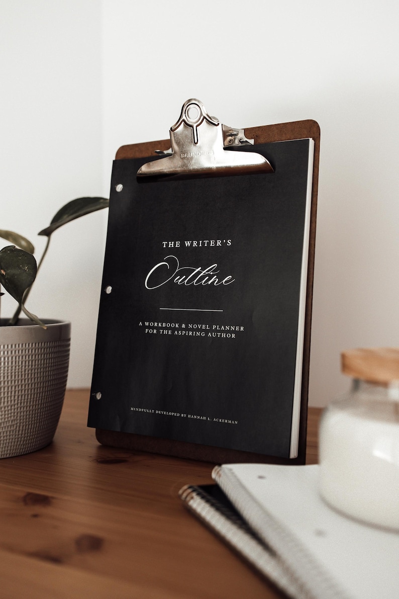 Może przedstawiać: Drewniana deska do pisania z metalowym klipsem trzymająca czarny notatnik zatytułowany "The Writer's Outline". Notatnik zawiera tekst "A Workbook & Novel Planner for the Aspiring Author". Deska do pisania spoczywa na drewnianej powierzchni.