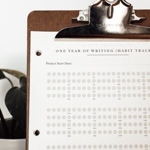 Może przedstawiać: Brązowa deska do pisania trzyma białą kartkę z napisem "ONE YEAR OF WRITING (HABIT TRACKER)". Papier ma siatkę k&oacute;ł do śledzenia postęp&oacute;w. W tle znajduje się roślina doniczkowa z ciemnozielonymi liśćmi.