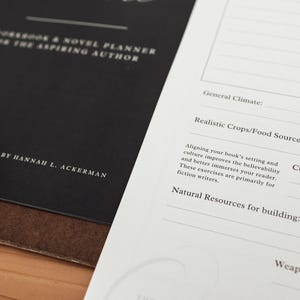 Może przedstawiać: Zeszyt ćwiczeń i planer powieści dla początkujących autor&oacute;w. Czarna okładka zawiera biały tekst, w tym "Opracowane przez Hannah L. Ackerman". Otwarta strona zawiera podpowiedzi dotyczące budowania świata.