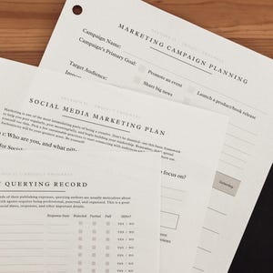 Może przedstawiać: Stos białych arkuszy papieru z nadrukowanym tekstem, w tym "Marketing Campaign Planning" i "Social Media Marketing Plan". Papiery mają jasnobeżowy odcień i są ułożone na drewnianej powierzchni.