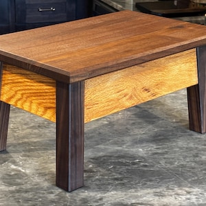 May include: A small wooden step stool featuring a dark brown rectangular top and four matching dark brown legs. The front panel is constructed from a contrasting light golden-brown wood with a visible grain pattern, set between the legs.