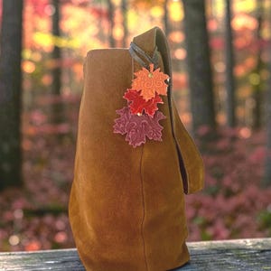May include: A brown suede shoulder bag featuring a decorative charm made of three stacked leather maple leaves in shades of orange, red, and deep burgundy. Each leaf has an embossed swirling pattern and is attached with a small metal rivet.