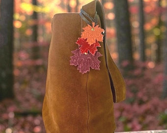May include: A brown suede shoulder bag featuring a decorative charm made of three stacked leather maple leaves in shades of orange, red, and deep burgundy. Each leaf has an embossed swirling pattern and is attached with a small metal rivet.