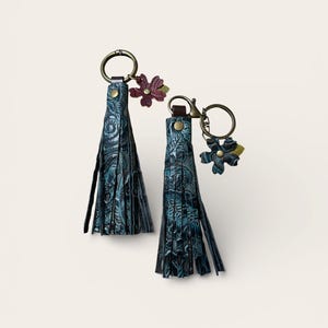 May include: Two teal leather tassel keyrings with floral accents. The tassels have a textured, embossed pattern and are attached to bronze-coloured key rings. One keyring has a burgundy flower, the other has a teal flower.