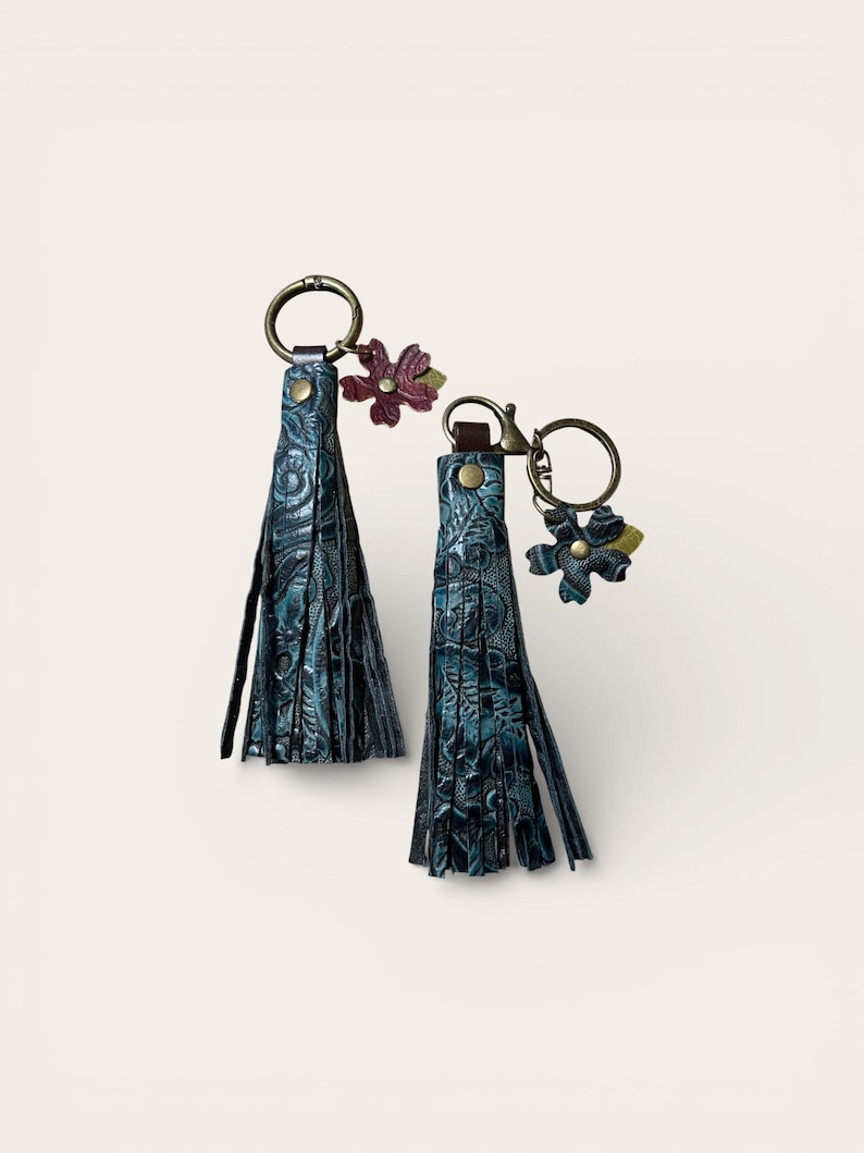 May include: Two teal leather tassel keyrings with floral accents. The tassels have a textured, embossed pattern and are attached to bronze-coloured key rings. One keyring has a burgundy flower, the other has a teal flower.