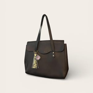 May include: A black leather tote bag with a flap closure and long handles. A decorative tassel with a floral accent hangs from the handle. The bag is a classic design, suitable for everyday use.