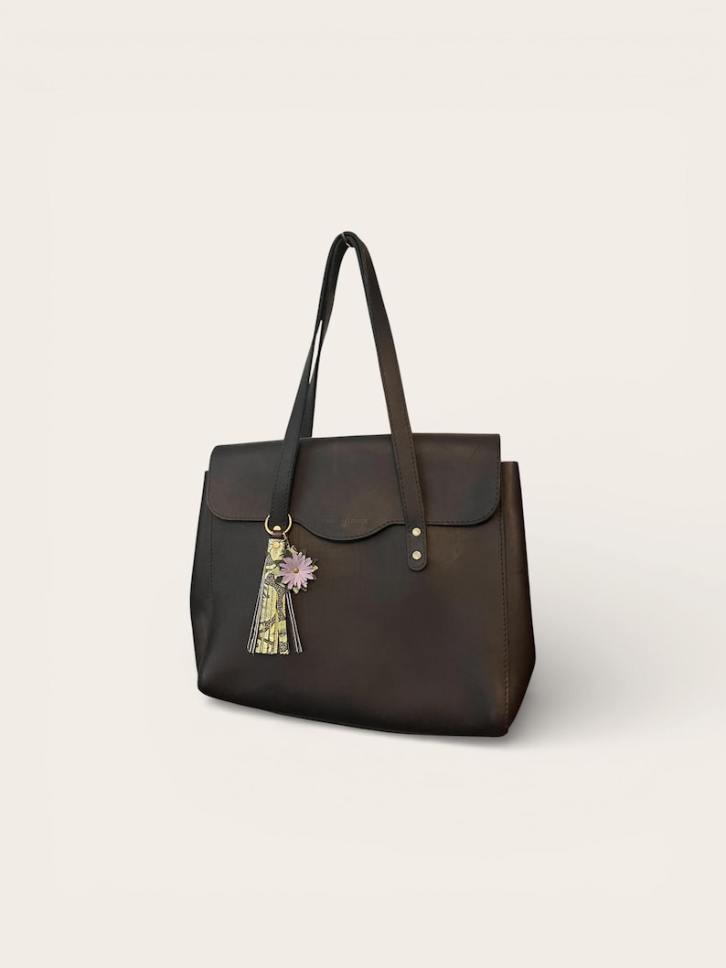 May include: A black leather tote bag with a flap closure and long handles. A decorative tassel with a floral accent hangs from the handle. The bag is a classic design, suitable for everyday use.
