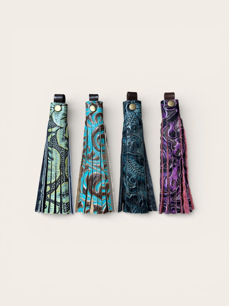 May include: Four leather tassels in a row, each with a different colour and pattern. The colours include green, turquoise, dark teal, and purple. Each tassel has a leather loop and a metal button.