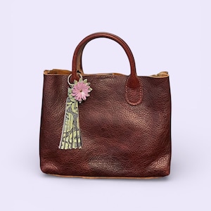May include: A brown leather tote bag with a textured finish and two handles. A pink flower and green tassel charm hangs from one handle. The bag has a square shape and a simple design.