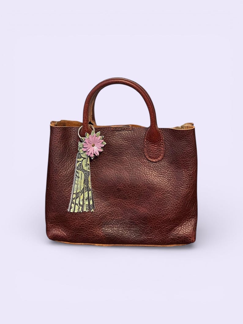 May include: A brown leather tote bag with a textured finish and two handles. A pink flower and green tassel charm hangs from one handle. The bag has a square shape and a simple design.