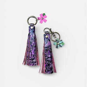 May include: Two purple leather tassel keyrings with floral accents. The tassels have a floral pattern and pink fringe. One keyring has a pink flower charm, the other a blue and green charm.