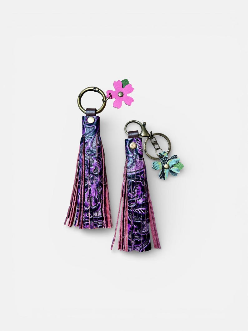 May include: Two purple leather tassel keyrings with floral accents. The tassels have a floral pattern and pink fringe. One keyring has a pink flower charm, the other a blue and green charm.