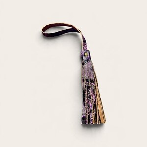 May include: A decorative tassel featuring a loop handle and multiple hanging strips of leather. The material displays a dark purple and black patterned design with a metallic gold-toned rivet securing the top of the fringe.