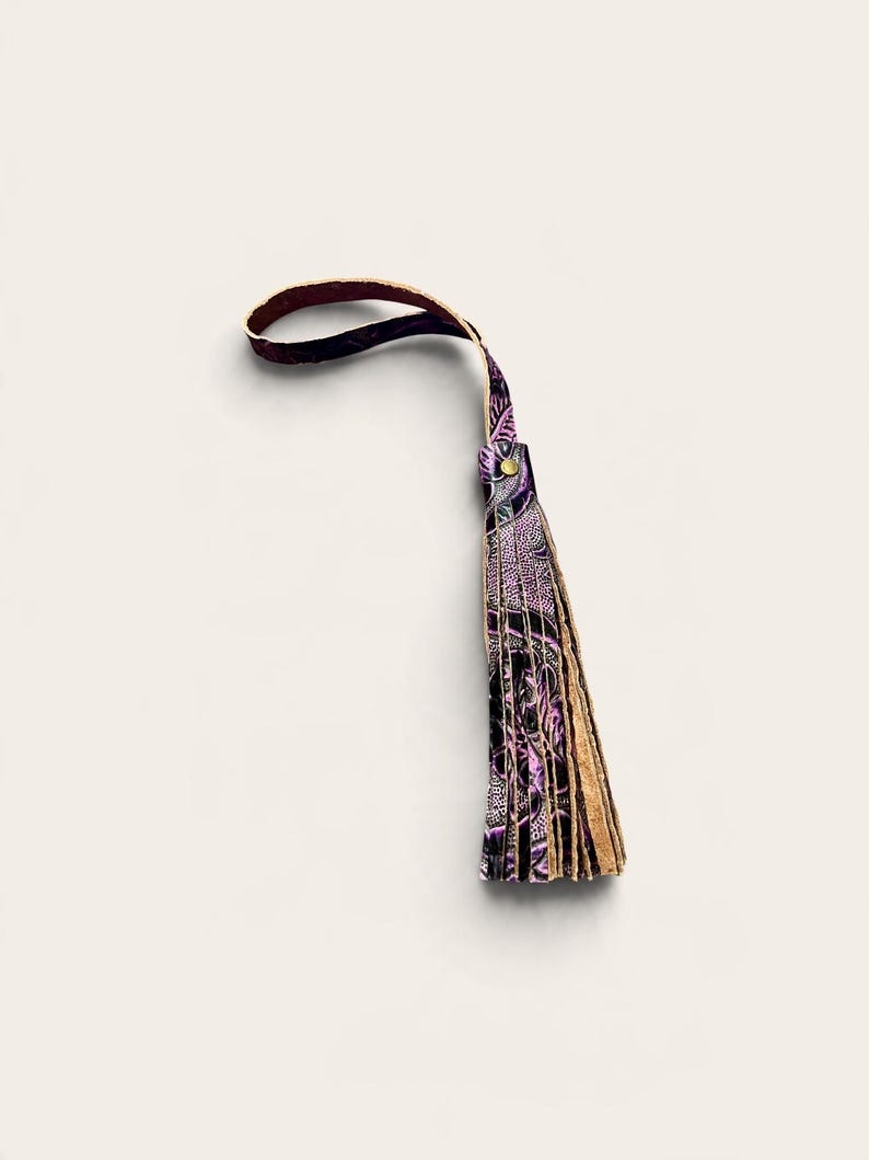 May include: A decorative tassel featuring a loop handle and multiple hanging strips of leather. The material displays a dark purple and black patterned design with a metallic gold-toned rivet securing the top of the fringe.