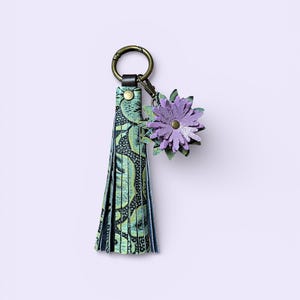 May include: A decorative keychain with a green and black patterned tassel and a purple flower charm. The keychain has a bronze-coloured ring and clasp. The flower charm is made of layered petals.