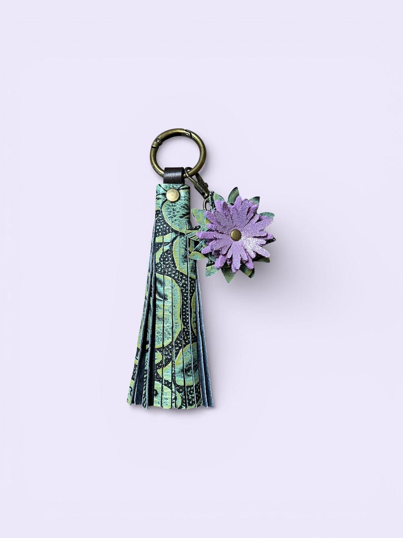 May include: A decorative keychain with a green and black patterned tassel and a purple flower charm. The keychain has a bronze-coloured ring and clasp. The flower charm is made of layered petals.