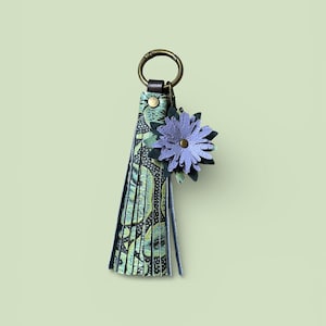 May include: A decorative keychain with a floral design. The keychain features a tassel with a green and black pattern, a metal ring, and a purple flower accent. The flower has a gold centre and is made of layered petals.